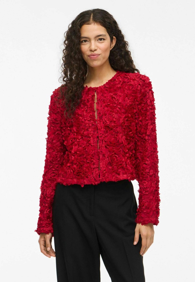 Veste courte à texture florale rouge, avec des manches longues, un col rond et une ouverture centrale. Présente des détails en pétales superposés et un ourlet sobre.