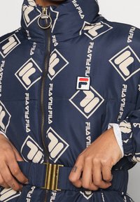 Granatowa puchówka z kremowym wzorem logo FILA, złotym zamkiem, wysokim kołnierzem, paskiem w talii i elastycznymi rękawami zapewniającymi dopasowany wygląd.
