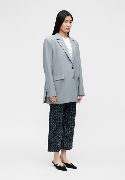 Γυναίκα που στέκεται φορώντας ένα ανοιχτό γκρι oversized σακάκι, λευκό τοπ, μαύρο και άσπρο παντελόνι με μοτίβο που κόβεται πάνω από τον αστράγαλο, και μαύρα μυτερά kitten heels.