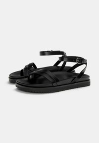 Sandales noires avec plusieurs brides croisées, boucle à la cheville, semelle plate en caoutchouc et finition en cuir lisse. Design minimaliste sans motifs.