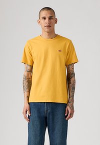 Man met getatoeëerde armen, die een geel kortshirt van Levi's en een blauwe spijkerbroek draagt, terwijl hij tegen een effen lichte achtergrond staat.