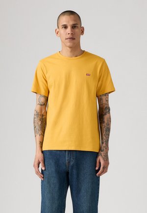 Man met getatoeëerde armen, die een geel kortshirt van Levi's en een blauwe spijkerbroek draagt, terwijl hij tegen een effen lichte achtergrond staat.