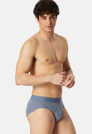 Junger Mann mit lockigem Haar, der graue Unterhosen trägt und vor einem schlichten weißen Hintergrund steht, blickt in die Kamera.