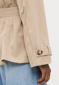 Beige jakke med folder i livet, med en knappestitt på mansjettene og glatt tekstur. Kombinert med lyseblå denim.