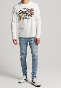Camisa blanca de manga larga con estampado gráfico, combinada con jeans desgastados de color azul claro y zapatillas blancas con detalles en negro.