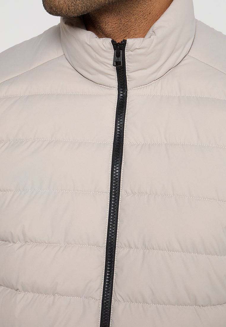 Hellbeige Pufferjacke mit hohem Kragen, horizontalen Nähten und einem dunklen Reißverschluss. Glatte, matte Textur. Keine sichtbaren Logos.