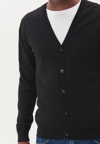 Černý button-up cardigan vyrobený z jemného materiálu, s designem do V, žebrovanými manžetami a šesti knoflíky vpředu. Nosič má na sobě bílou košili.