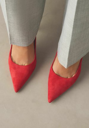 Füße, die leuchtend rote spitze Schuhe tragen, kombiniert mit hellgrauen Hosen auf einem neutralen beigen Boden.