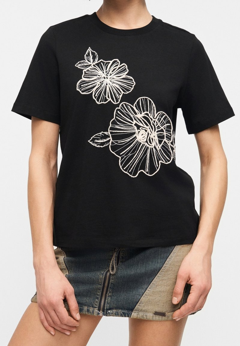 T-shirt noir à manches courtes avec de grandes fleurs blanches brodées, associé à une mini-jupe en denim color block avec une fermeture éclair à l'avant.