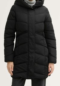 Abrigo acolchado negro con capucha, usado sobre un suéter de cuello alto blanco, con bolsillos laterales con cremallera y mangas largas.