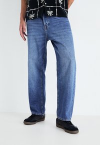 Jeans de denim azul con un corte relajado, con un sutil desgastado y detalles de costura. Combinados con zapatillas negras.