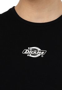 Svart bomullströja med rund halsringning; har en vit logotyp "Dickies" med en cirkulär design ovanför texten.