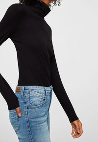 Top negro de manga larga y cuello alto con un diseño ajustado, acompañado de jeans de tiro alto en un tono azul claro que cuentan con un parche de piel en la parte trasera.