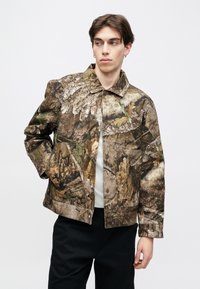 Μπουφάν με καμουφλάζ σε καφέ αποχρώσεις, κατασκευασμένο από υφή υφάσματος, με γιακά, κουμπιά τύπου ανοιγόμενο και εμφανείς λεπτομέρειες ραφής.