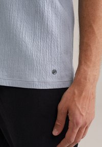 Világoskék, textúrázott ing hullámos mintával, rövid ujjakkal és logós részlettel az alsó szegélyen, fekete szövetnadrággal párosítva.