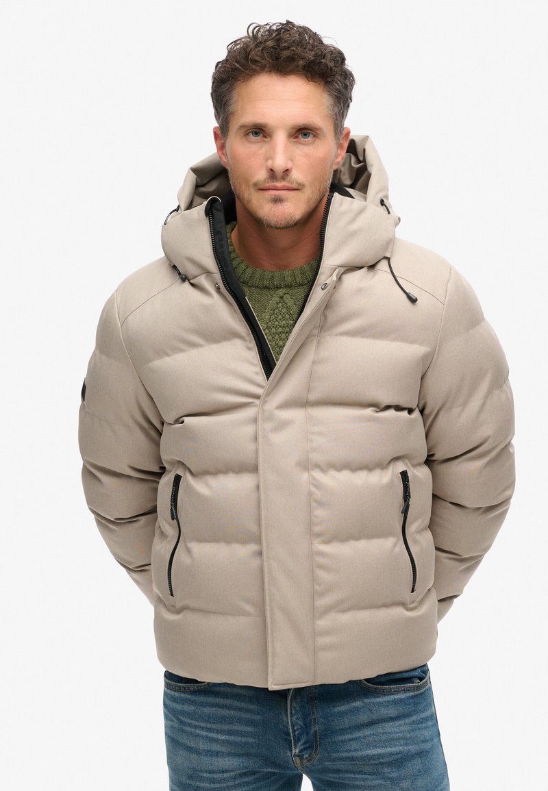 Beige pufferjack met capuchon, met een gequiltde textuur, ritszakken aan de zijkant en een hoge kraag. Draagt over een groene trui en blauwe jeans.