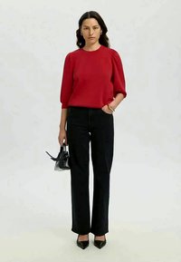Mulher a usar uma blusa vermelha e calças pretas, segurando uma pequena bolsa preta, de pé contra um fundo claro e liso.