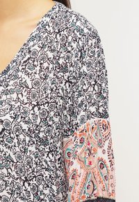 Blusa estampada con un diseño floral en blanco y negro, con acentos en turquesa y un estilo paisley naranja en las mangas.