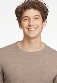 Ljusbrun stickad tröja med rund halsringning och subtil struktur. Har en liten broderad logotyp på vänstra bröstområdet.