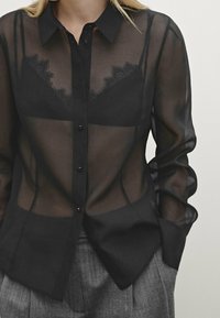 Ohut musta paita, jossa on muotoiltu kaulus, etupainonapit ja pitsikoristelu kankaan alla. Pitkät hihat pyöreillä cuffeilla.