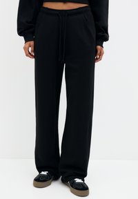 Pantalons de jogging larges et noirs avec une taille élastique et un cordon de serrage. Comprend deux poches latérales et un tissu doux et texturé. Associés à des baskets noires.