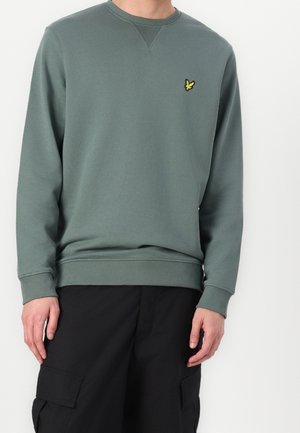 Homme portant un sweat-shirt vert sourd à col rond avec un petit logo d'oiseau jaune sur la poitrine et un pantalon cargo noir, debout devant un fond blanc.