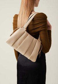 Beige gefaltete Handtasche mit glattem Ledertextur, goldenen Hardware-Akzenten und verstellbaren Riemen. Das Design präsentiert eine strukturierte, moderne Silhouette.