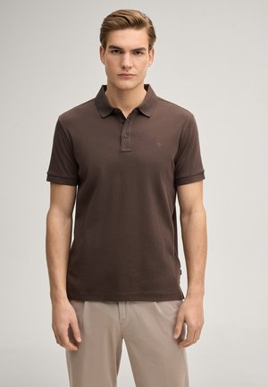 Junger Mann steht, trägt ein braunes kurzärmeliges Polohemd und beige Hosen, blickt nach vorne vor einem schlichten Hintergrund.