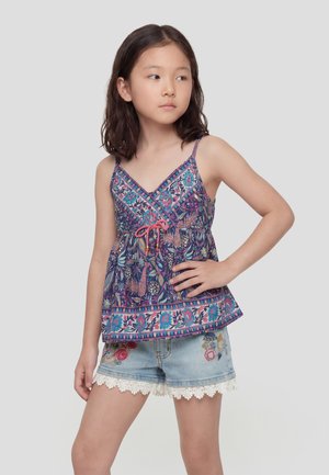 Joven de pie con la mano en la cadera, vistiendo una blusa sin mangas azul con flores y pantalones cortos de mezclilla con ribete de encaje sobre un fondo gris claro.