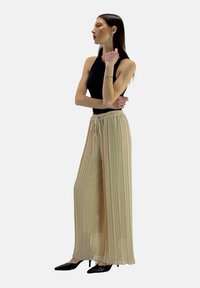 Beige, geplooide wijde broek met een elastische tailleband en trekkoord, gecombineerd met een aansluitend zwart mouwloos topje en zwarte hakken.
