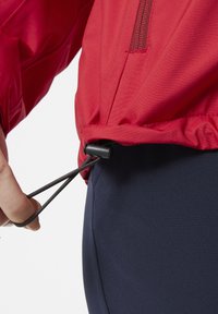 Rode jas met een verstrakkende toggle aan de onderrand, voorzien van een horizontale ritszak. Achtergrond is donkerblauwe stof, wat suggereert dat het om buitenkleding gaat.