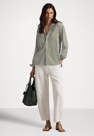 Γυναίκα με φαρδιά ριγέ πουκάμισο με κουμπιά, λευκό παντελόνι, καφέ σανδάλια, κρατώντας σκούρα πράσινη τσάντα, στέκεται μπροστά σε απλό φόντο ανοιχτού χρώματος.