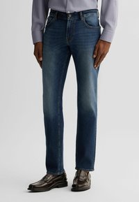 Jeans in denim blu con una forma aderente e una finitura leggermente sbiadita. Presentano cinque tasche e una chiusura con bottoni, indossati con scarpe in pelle scura.