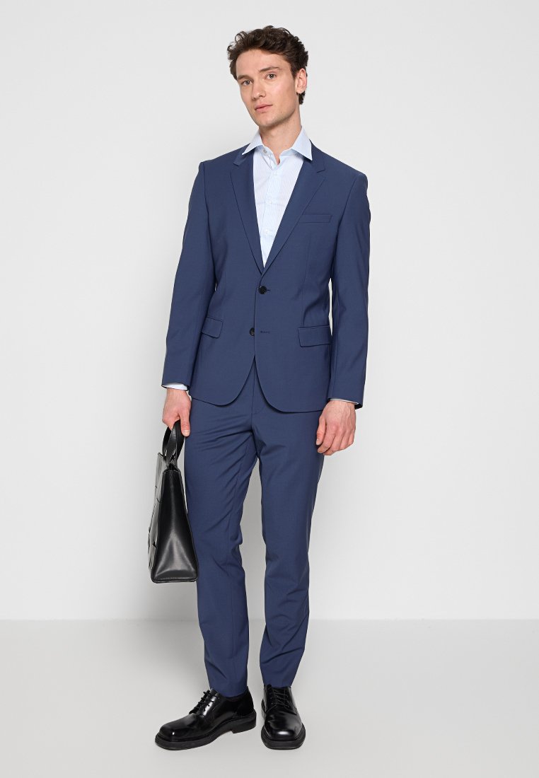 Uomo in abito blu navy e camicia azzurro chiaro che tiene una valigetta in pelle nera, in piedi davanti a uno sfondo bianco.