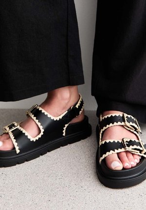 Zwarte leren sandalen met crèmekleurige zigzagaccenten, voorzien van twee verstelbare riemen en een dikke rubberen zool. Textuurafwerking en open ontwerp.