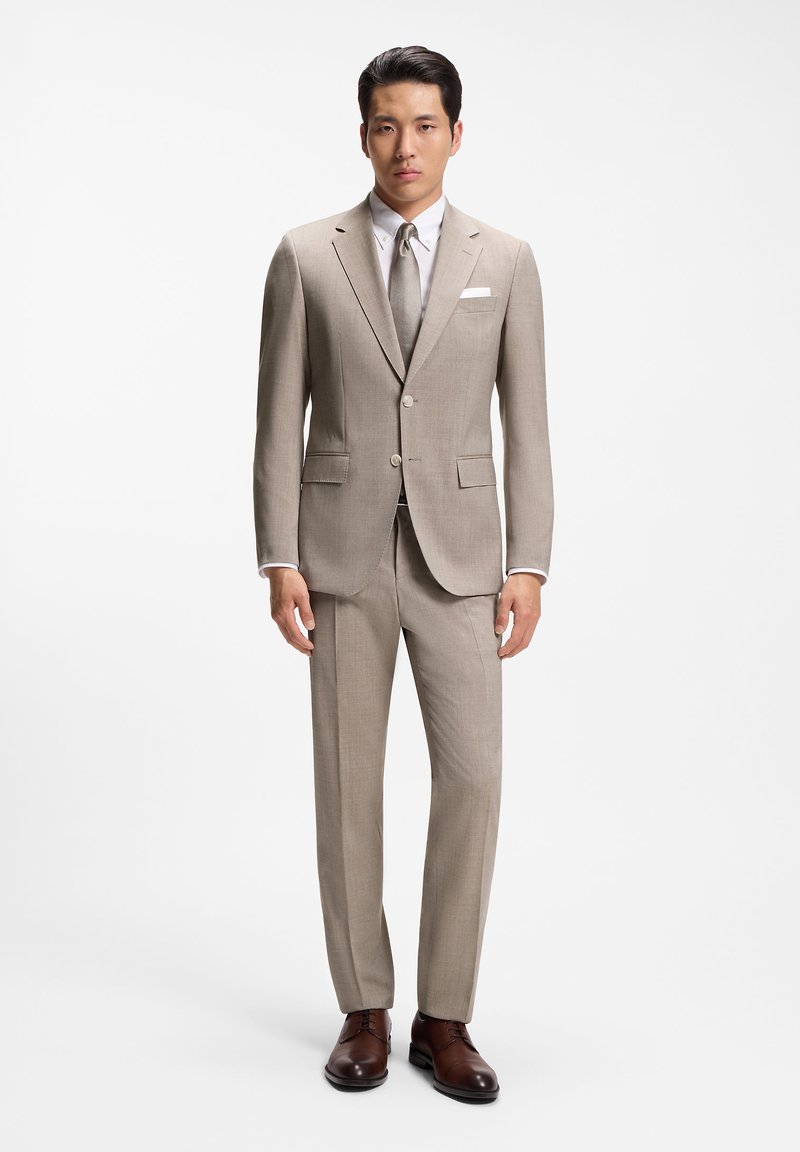 Un homme se tient debout portant un costume beige à deux boutons, une chemise blanche, une cravate claire, un mouchoir blanc dans la poche et des chaussures habillées en cuir marron sur un fond blanc.