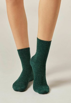 Personne portant des chaussettes côtelées de couleur vert foncé avec une texture mouchetée, se tenant sur une surface de couleur claire.