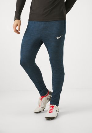 Mężczyzna w ciemnoniebieskich legginsach Nike i białych butach piłkarskich Nike z czerwonymi sznurowadłami, stojący na jednolitym białym tle.