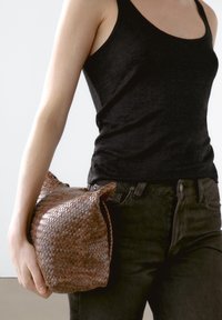 Vävd brun väska med en mjuk, rundad form, hålls i en hand. Materialet verkar vara texturerat med ett subtilt mönster. Mörkt linne och jeans syns.