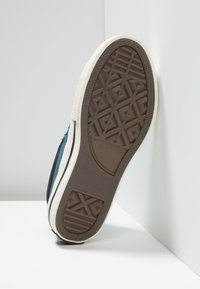 Hnědá gumová podrážka s texturovaným vzorem; bílá střední podrážka; modrý a černý textilní svršek; značka Converse All Star viditelná na podrážce.