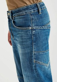 Zijaanzicht van een persoon die losse blauwe denim jeans draagt met een grote zijvak en een ingestopte bruine shirt.