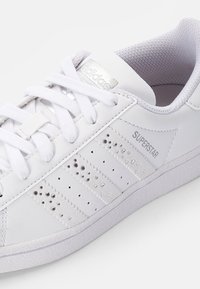 White Adidas Superstar sneakers featuring a smooth leather upper, three striped design with stud accents, and a textured rubber sole.