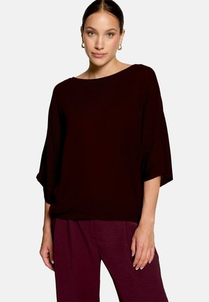 Femme aux cheveux attachés portant des boucles d'oreilles anneaux en or, un haut bordeaux foncé ample et un pantalon assorti sur un fond clair uni.