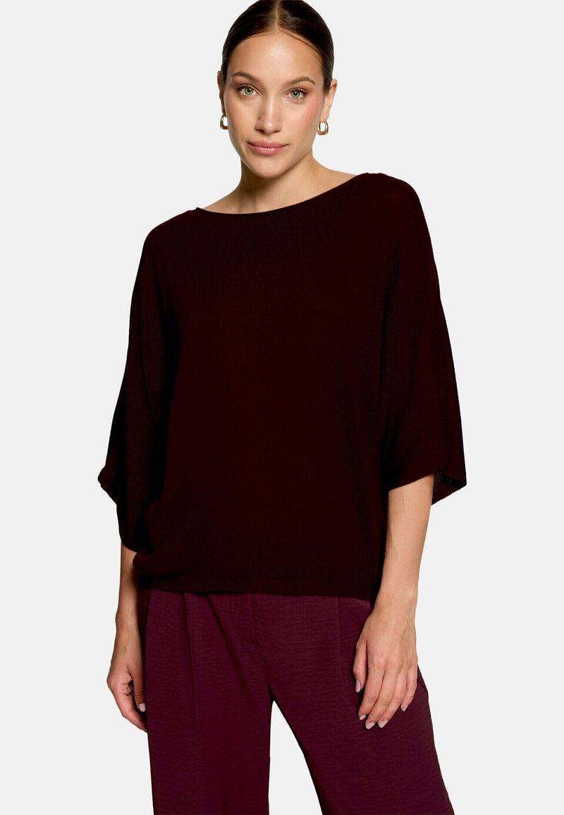 Femme aux cheveux attachés portant des boucles d'oreilles anneaux en or, un haut bordeaux foncé ample et un pantalon assorti sur un fond clair uni.
