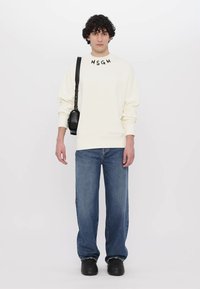 Sweatshirt crème avec le logo "MSGM", associé à un jean large bleu. Comprend un sac à bandoulière noir et des chaussures noires. Fond uni.