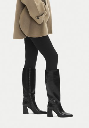 Donna in piedi di lato, indossa stivali neri in pelle con tacco alto fino al ginocchio, leggings neri e un cappotto beige con polsini abbottonati.