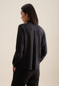 Blusa nera a maniche lunghe con taglio dritto, dettaglio a piega sul retro e texture morbida. Abbinata a pantaloni neri.