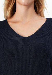 Prim-plan al unei persoane care poartă un pulover V cu striații de culoare albastru închis, care dezvăluie gâtul, clavicula și o parte din părul lung blond.