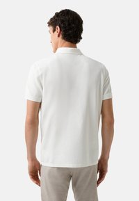 Hvid polo skjorte med en klassisk krave, korte ærmer og en glat tekstur, set bagfra, iført lyse bukser.