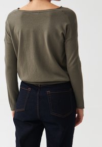 Femme portant un haut à manches longues vert olive et un jean bleu foncé, vue de dos contre un fond clair uni.